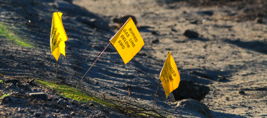Three small yellow warning flags planted in the ground along a dirt surface. The flags are labeled “Buried Gas Line Below SWG,” marking the location of underground utilities.