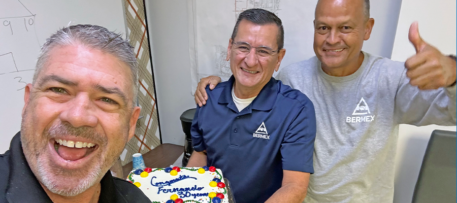 Three smiling men stand close together celebrating a work anniversary. The man on the left takes a cheerful selfie, while the man in the center holds a decorated sheet cake that reads “Congratulations… 30 years.” The man on the right gives a thumbs-up. Two of the men wear shirts with the Bermex logo. They appear to be in an office setting with charts and papers on the wall behind them.
