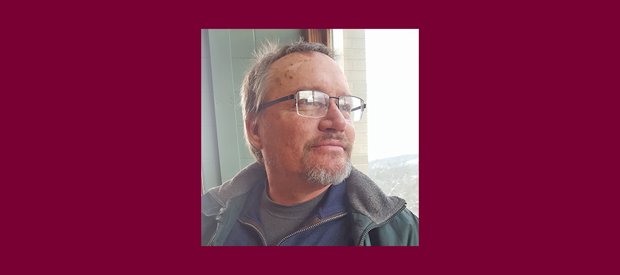 A man with short gray hair, glasses, and a trimmed beard looks out a window with soft daylight on his face.